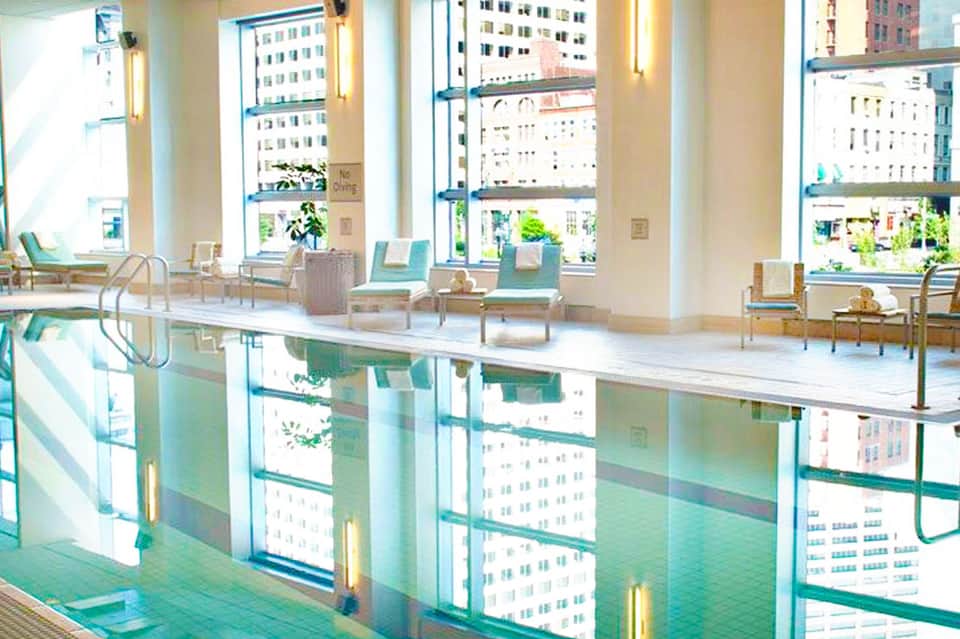 Indoor lap pool with floor-to-ceiling windows overlooking a city skyline, with lounge chairs nearby