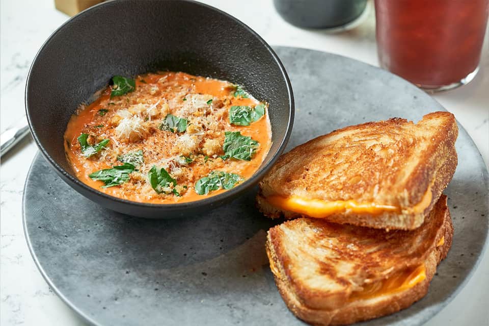 Gourmet tomato soup in black bowl with grilled cheese sandwich and red beverage on gray plate