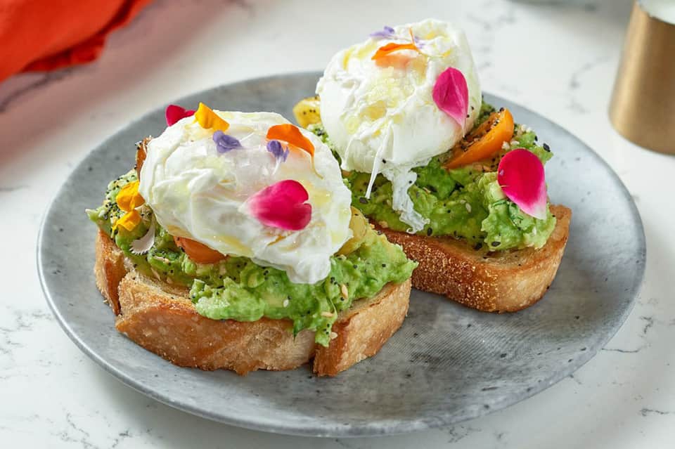 Gourmet avocado toast with poached eggs and colorful edible flowers on gray plate