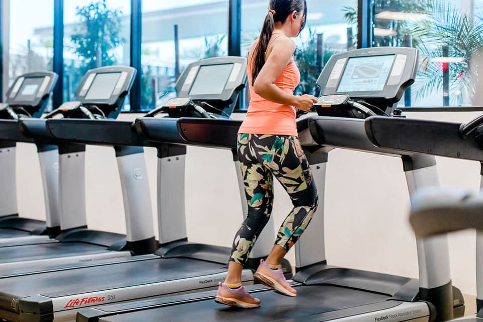 Woman running on treadmill in bright fitness center with floor-to-ceiling windows and ocean views