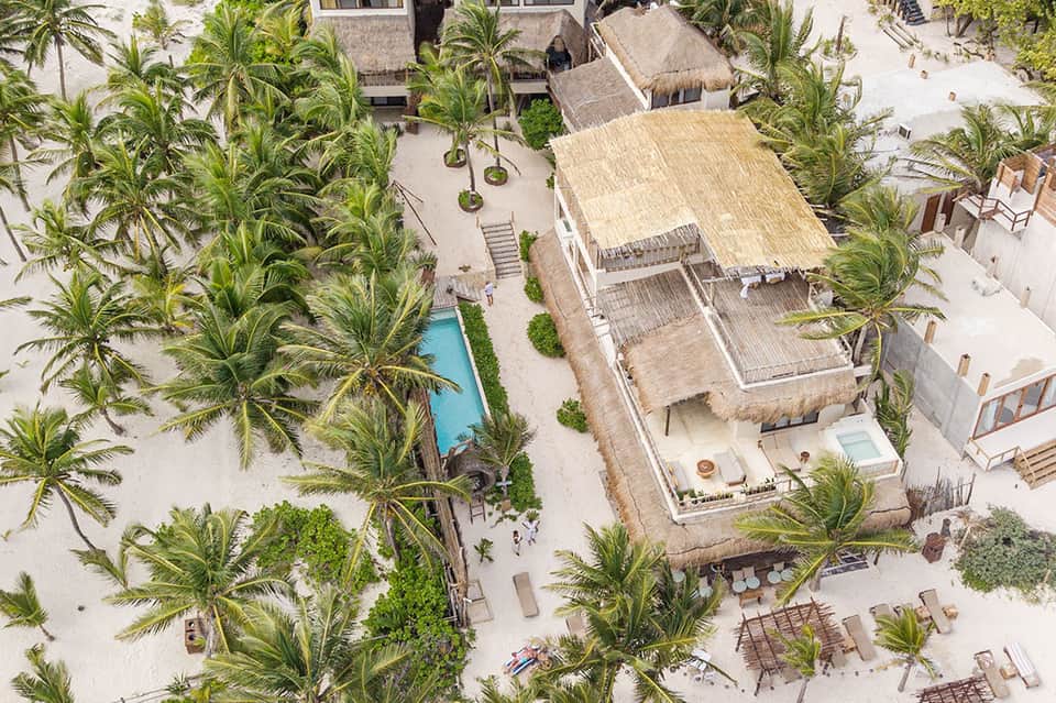 Aerial resort view with thatched-roof structures, white sand, palm trees, and small pool surrounded by lounge chairs