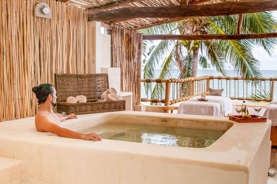 Guest relaxing in outdoor soaking tub on beachfront spa terrace with ocean views and tropical furnishings