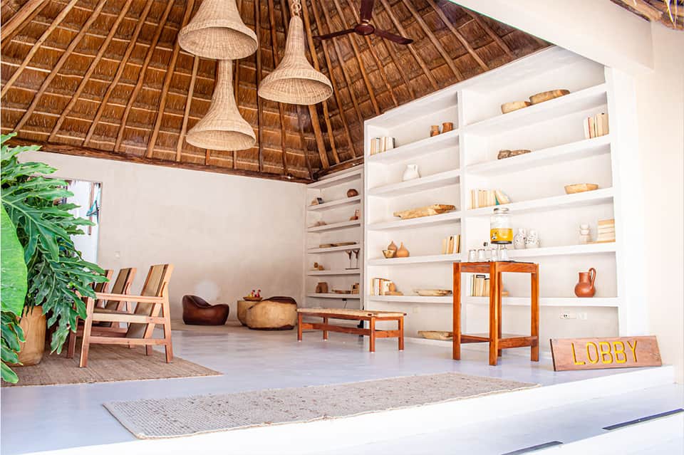 Modern resort lobby with thatched ceiling, white shelving, wooden furniture, and tropical plants
