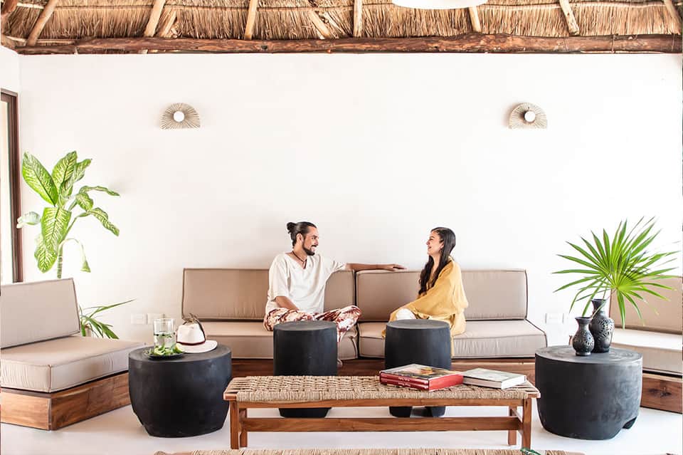 Two guests lounging on neutral sofa beneath thatched roof with plants and modern decor