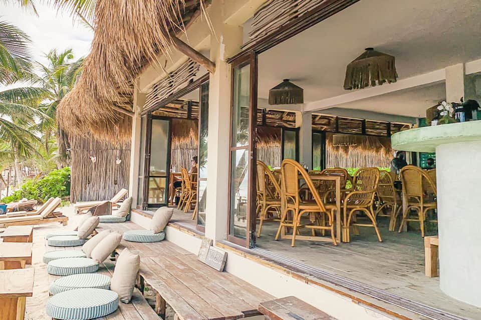 Tropical beachfront lounge with thatched umbrellas, wooden deck, cushioned seating, and ocean view through glass doors