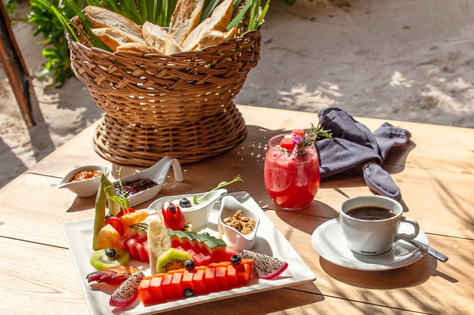 Beachside breakfast setup with fresh fruit plate, coffee, and tropical drink on wooden table