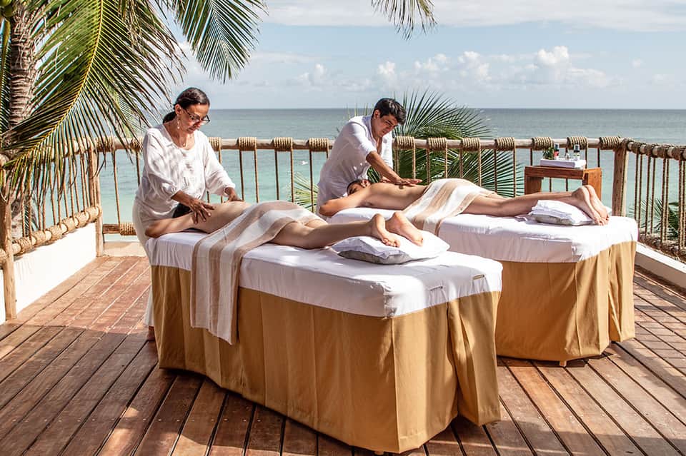 Beachfront spa treatment with two massage therapists working on clients overlooking ocean with palm fronds