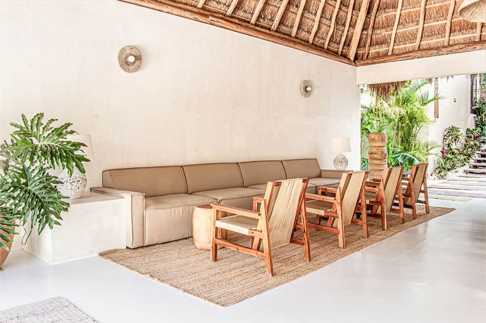 Tropical lounge with high thatched ceiling, taupe sofa, wooden chairs, and plants