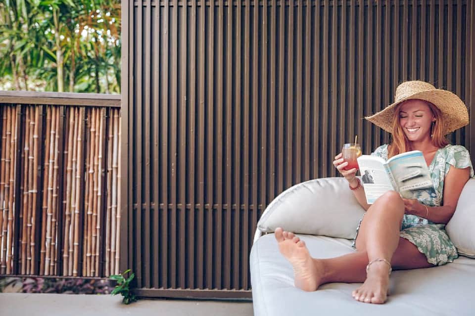 Woman relaxing on white lounge chair reading book, wearing straw hat near black fence