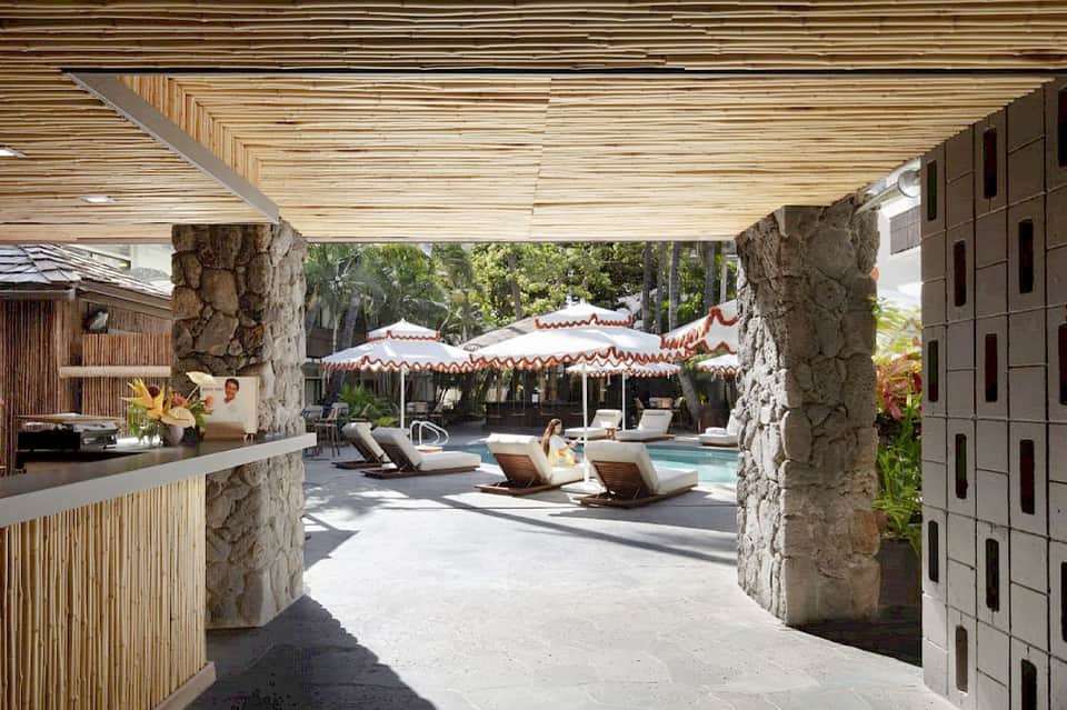 Resort pool deck with red and white umbrellas, loungers, stone walls, bar, and tropical landscaping