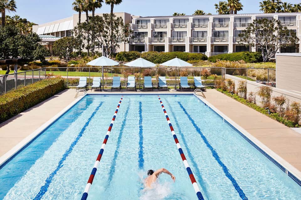 Olympic-length lap pool with lane dividers and lounge chairs under white umbrellas, modern hotel building in background