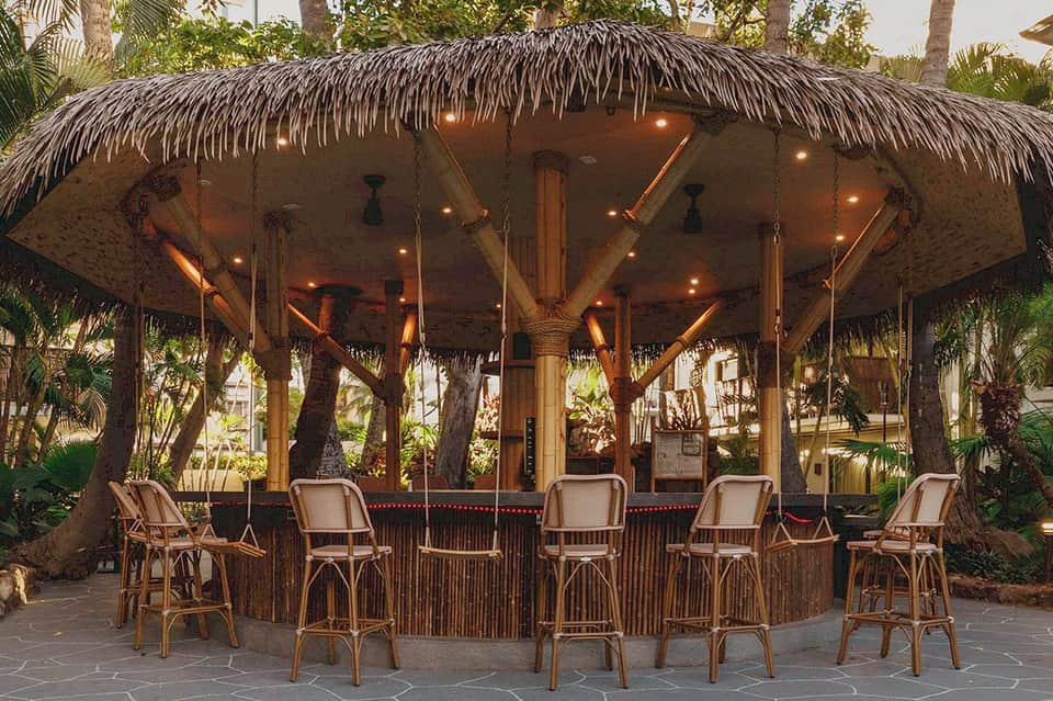Beachfront tiki bar with thatched roof, wooden railings, woven bar stools, and warm lighting