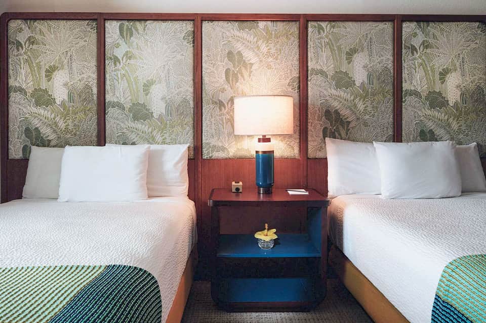 Hotel bedroom with two queen beds, botanical headboard panels, and blue accent furniture