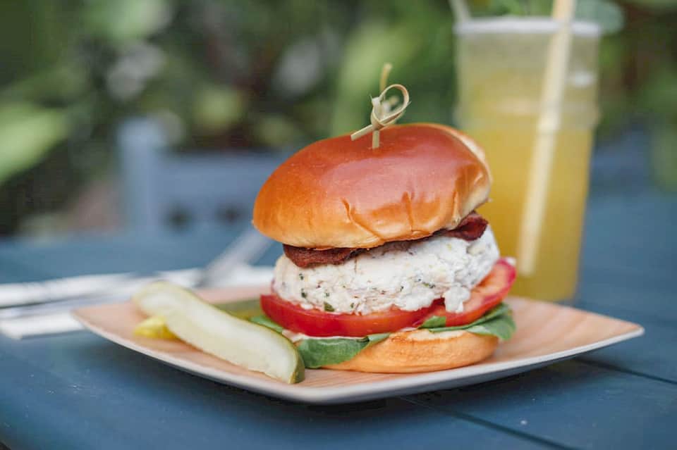 Gourmet burger with bacon, herb spread, tomato, and cucumber served with beverage outdoors