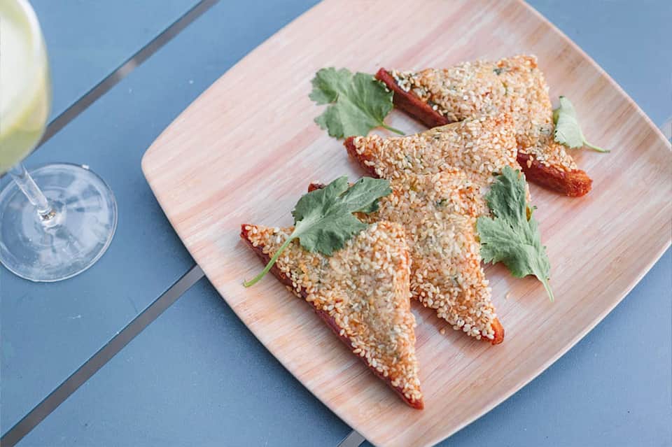 Gourmet appetizers with sesame coating and herbs on white wooden board next to cocktails