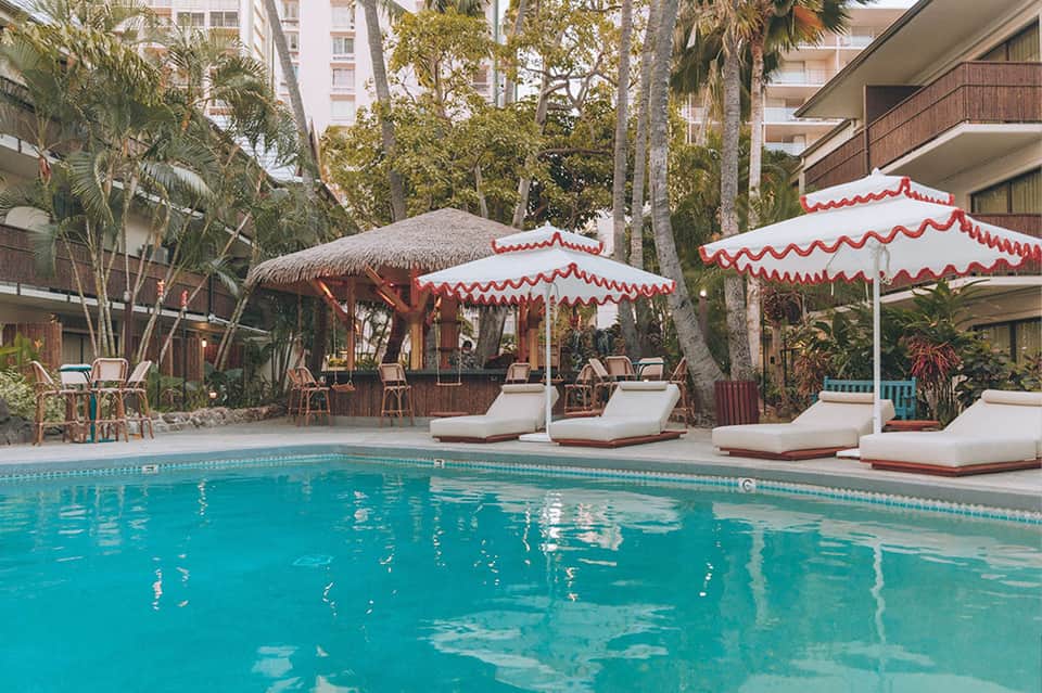 Resort pool with turquoise water, red and white umbrellas, white lounge chairs, palm trees, and tropical landscaping