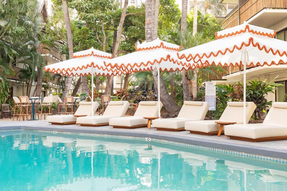Resort pool with orange and white striped umbrellas, cushioned loungers, and palm trees