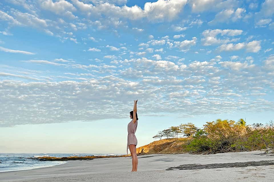 Woman in white dress standing on beach with arms raised under blue sky and clouds
