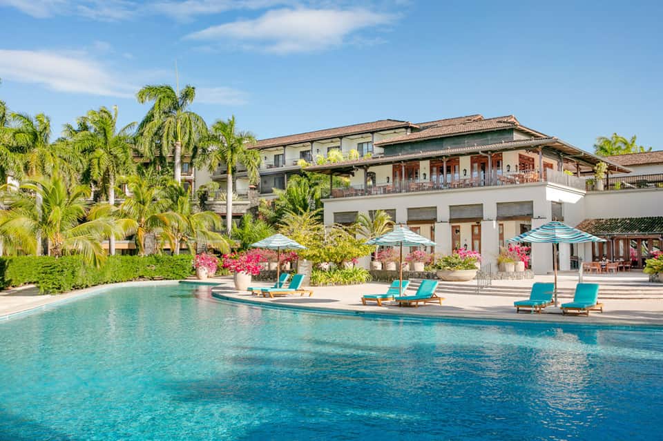 Luxury resort pool surrounded by palm trees, turquoise loungers, and Mediterranean architecture