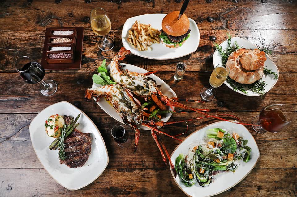 Fine dining table spread with lobster, burger, croissants, steak, and salad with wine on rustic wood