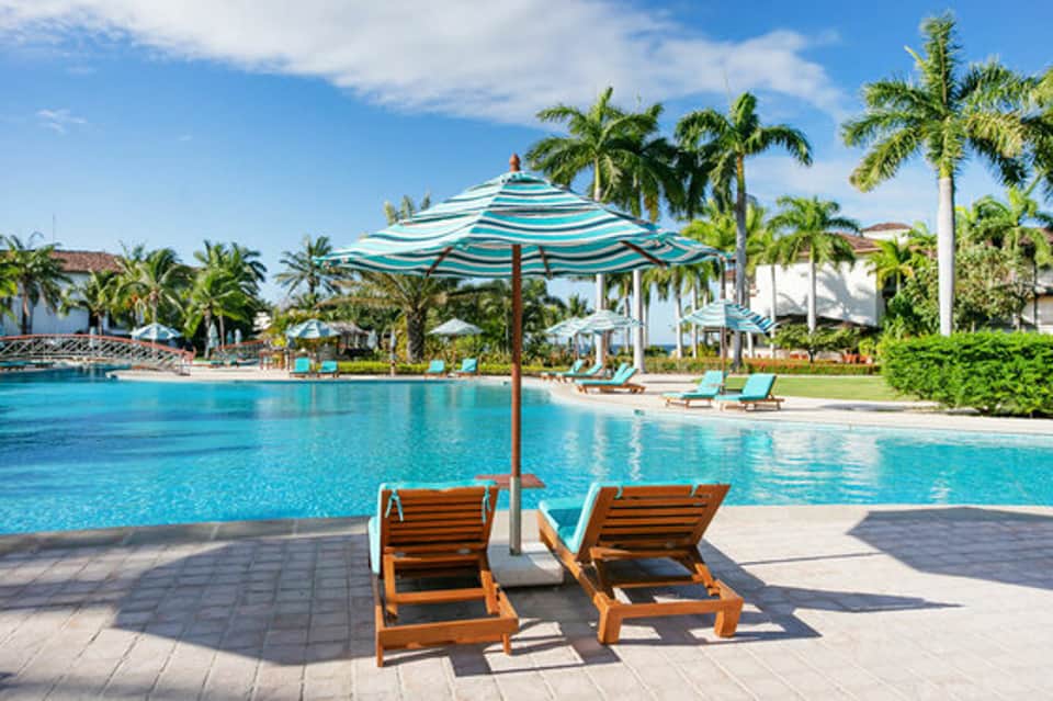 Beachfront resort pool with striped umbrella, lounge chairs, and palm trees