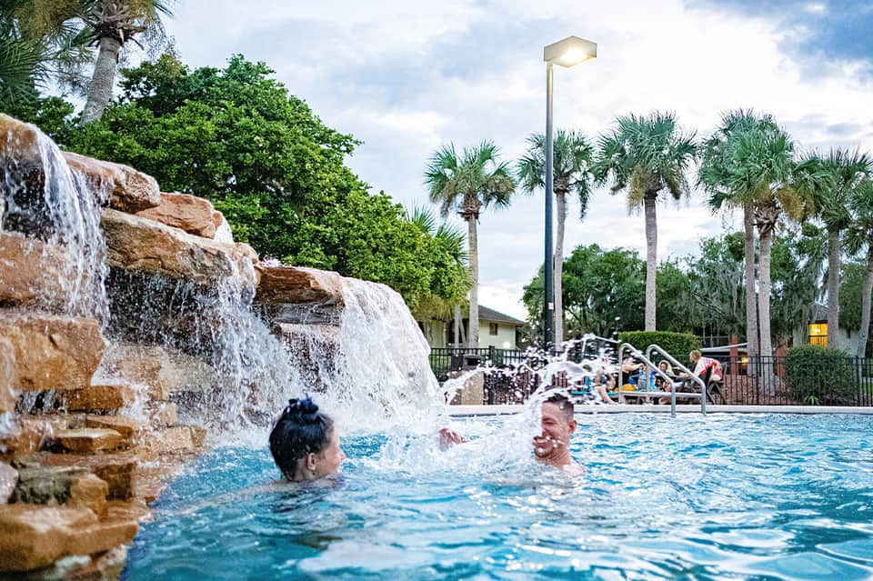 Resort pool with waterfall feature, palm trees, and guests swimming on sunny day