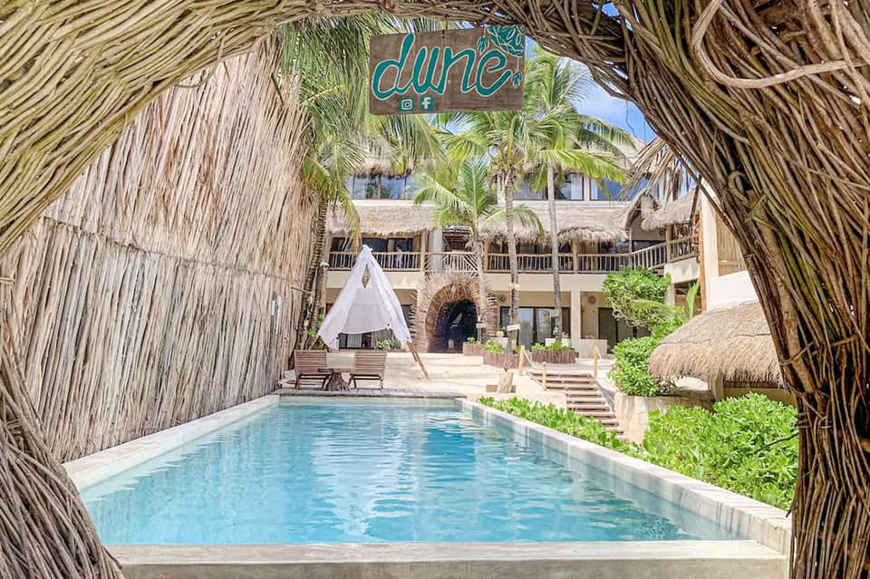 Thatched-roof covered pool pathway with turquoise water and resort building beyond