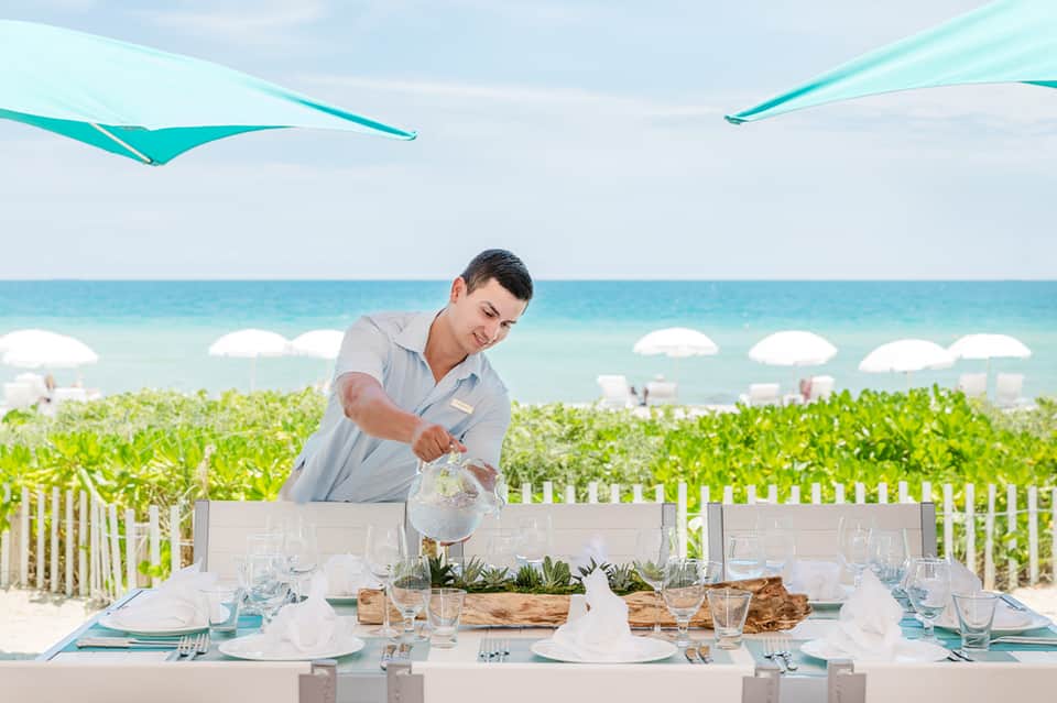 Server pouring water at beachfront dining table with turquoise umbrellas