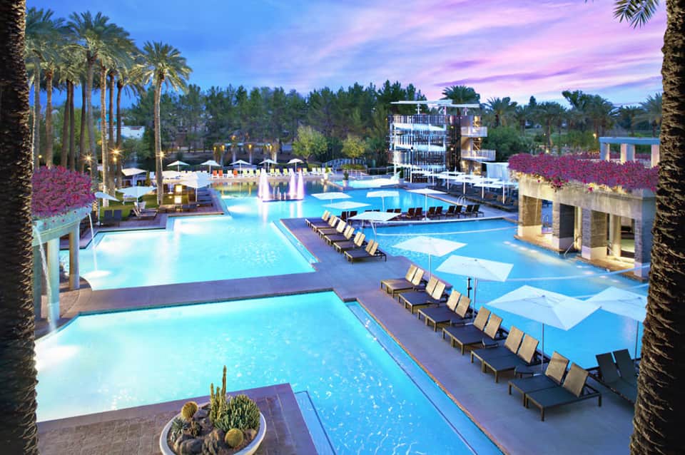 Expansive resort pool complex at dusk with palm trees, lounge chairs, and illuminated water features