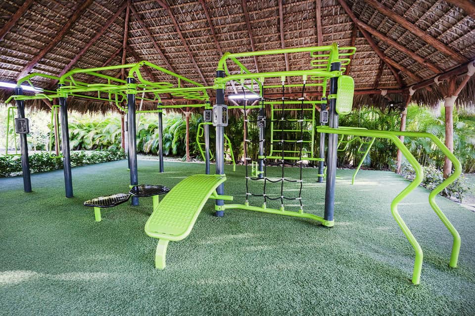 Bright green outdoor fitness equipment under thatched roof with tropical plants and landscaping