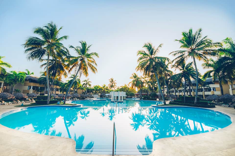 Large turquoise resort pool surrounded by palm trees and beige multi-story hotel buildings