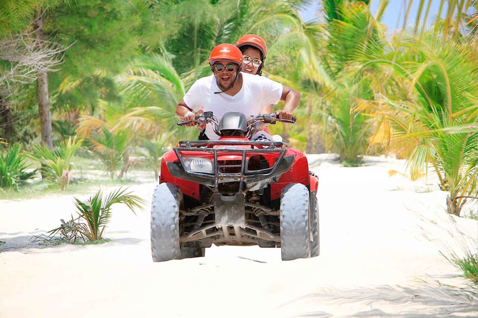 Two people riding a red ATV on a sandy beach lined with palm trees
