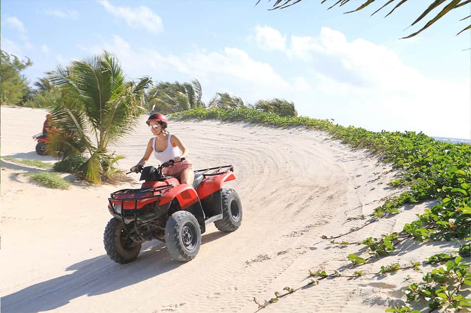 Woman riding red ATV along sandy beach with palm trees and coastal vegetation in background