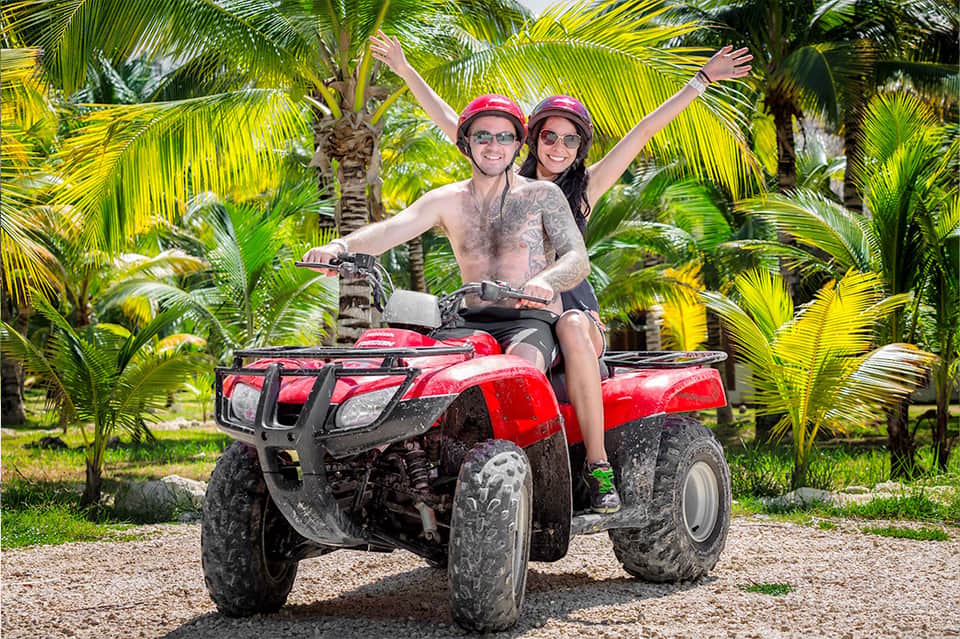 Couple on red ATV in tropical palm tree setting with arms raised in celebration