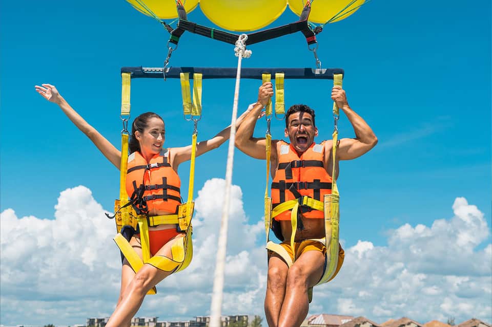 Two people parasailing over blue water wearing life vests attached to yellow parachute
