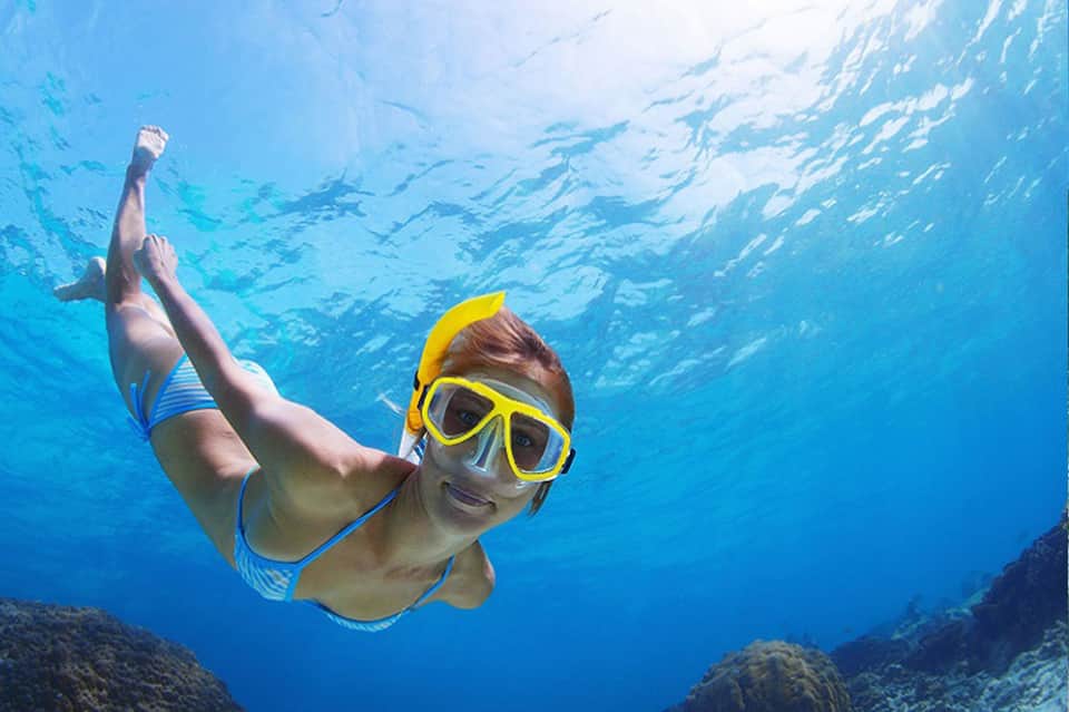 Snorkeler in yellow goggles underwater with coral reef, school of fish and sunlight streaming above