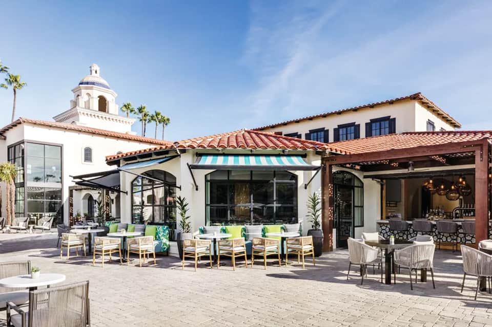 Spanish-style resort courtyard with white buildings, blue awnings, dining areas, and palm trees