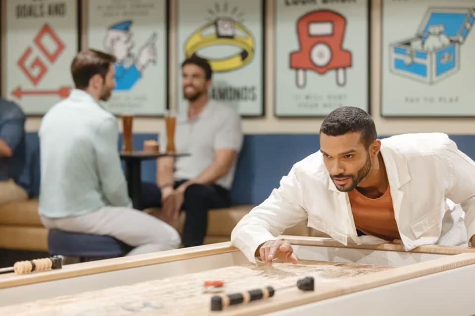 Man playing shuffleboard in rec room with vintage board game posters