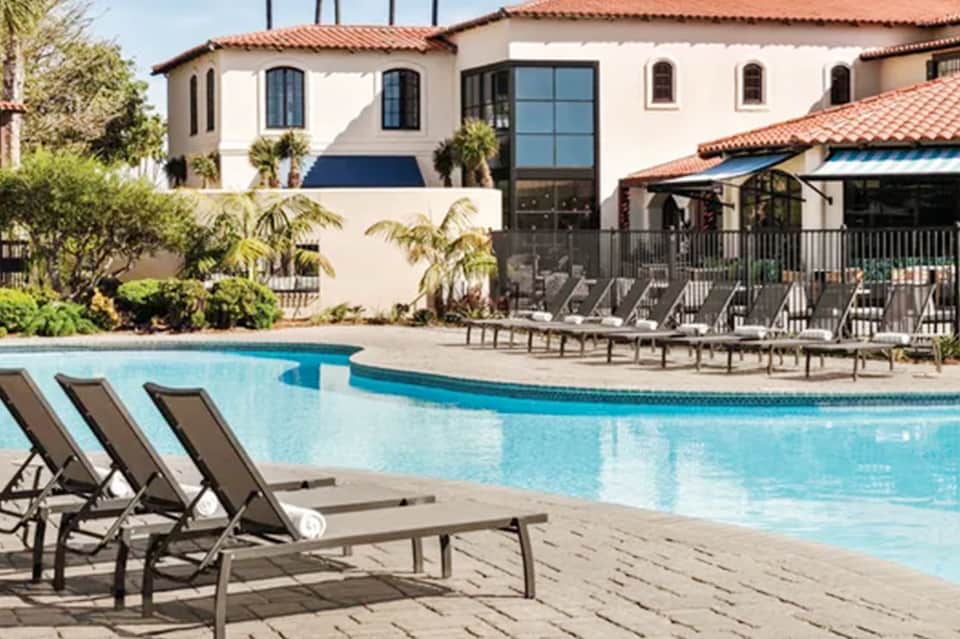 Spanish-style resort pool with lounge chairs, white building, palm trees, and covered patio area