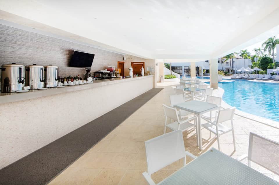 Indoor pool bar with white furniture, coffee machines, and columns overlooking resort swimming pool