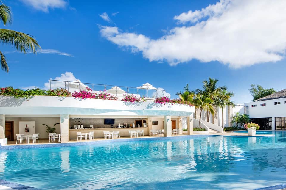 Resort swimming pool with white cabanas, bar, tropical landscaping, and mountain backdrop