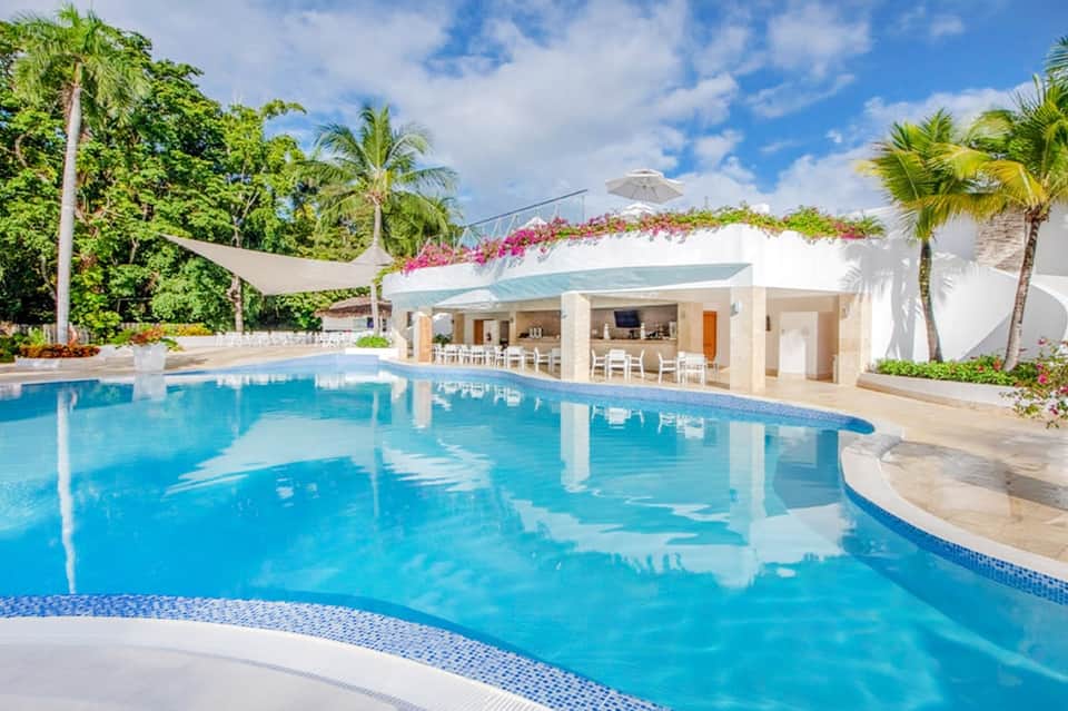 Curved resort pool with turquoise water, white lounge structure, pink flowers, and palm trees