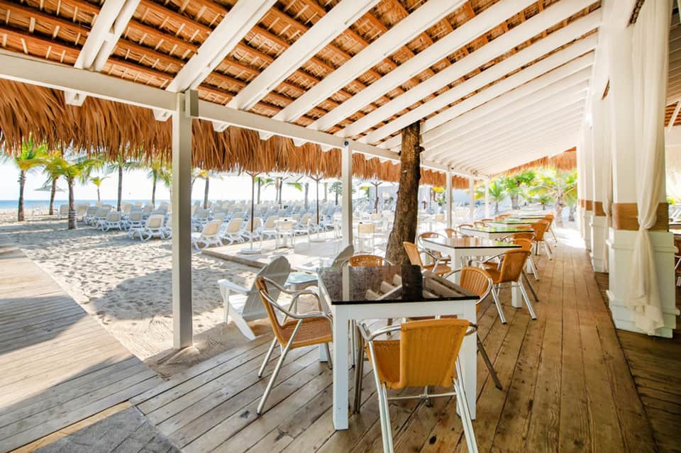 Beachfront dining pavilion with thatch roof, wooden tables, chairs, and direct beach access