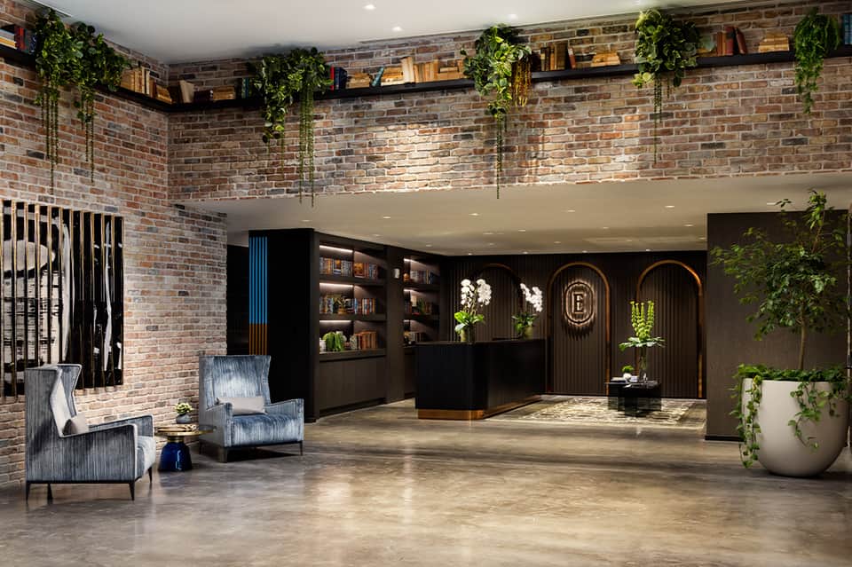 Contemporary hotel lobby with exposed brick walls, cascading plants, black reception desk, and modern furnishings