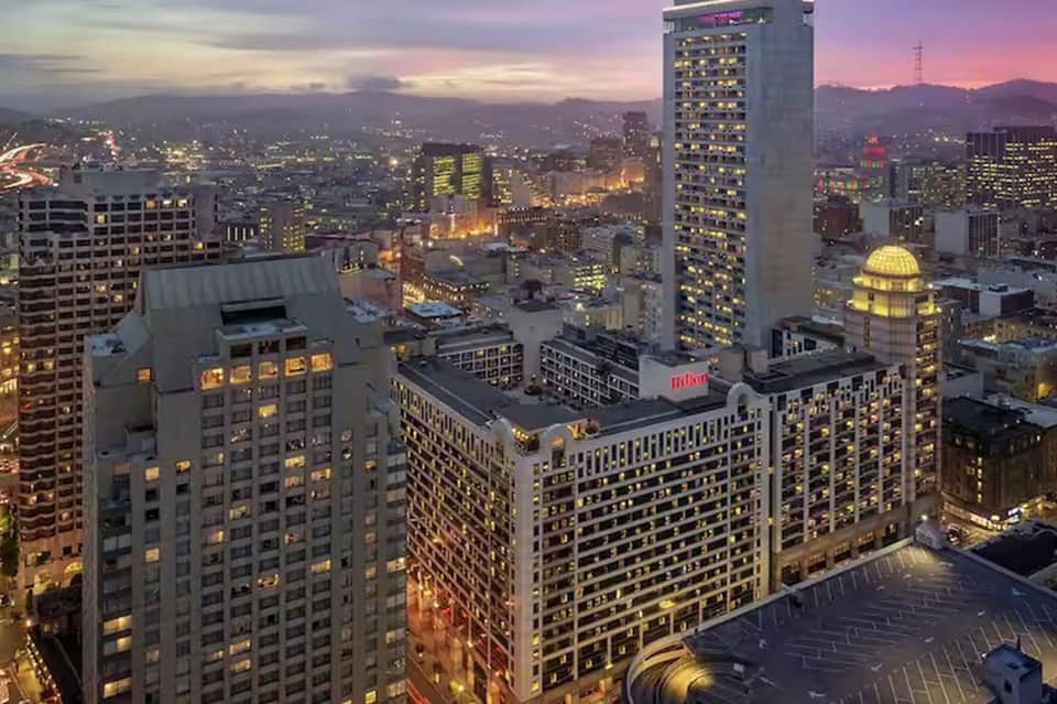 Urban cityscape at dusk with illuminated office buildings, dome structure, and mountain backdrop