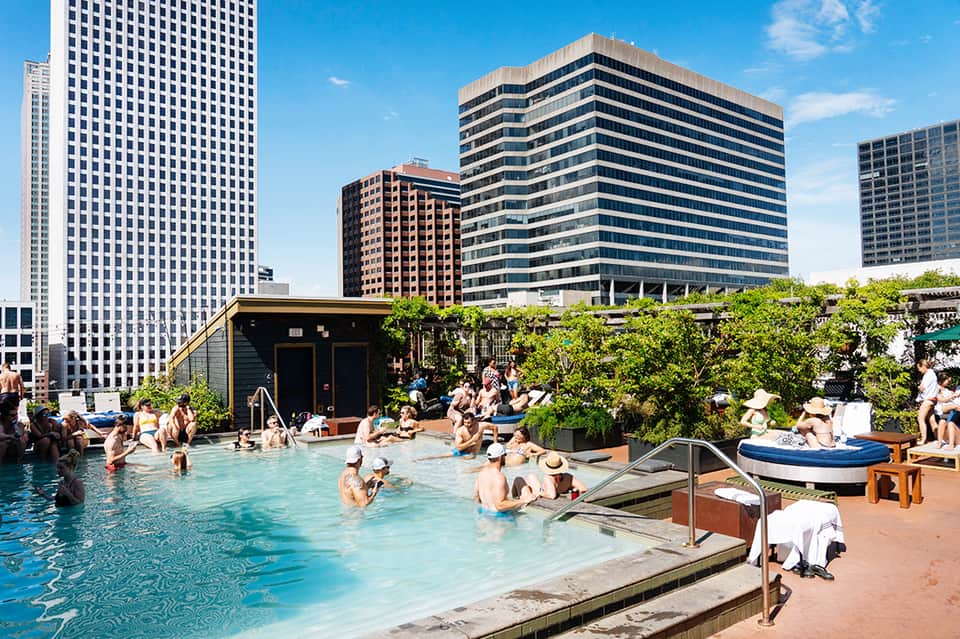 Rooftop pool at urban hotel with skyline views, guests swimming, lounge areas, and greenery