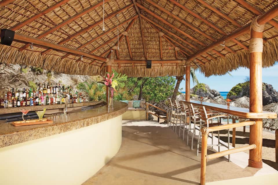 Thatched-roof beach bar with wooden details, bar counter, tropical flowers, and ocean views