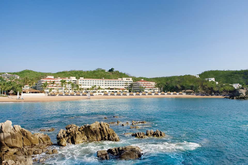 Beachfront resort viewed from ocean with rocky outcropping, sandy shore, and multi-story hotel buildings