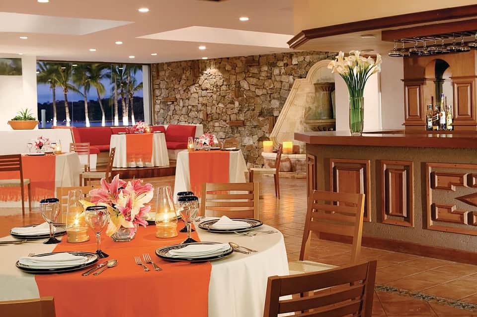 Elegant dining room with coral tablecloths, round tables, stone wall, and ocean view through windows