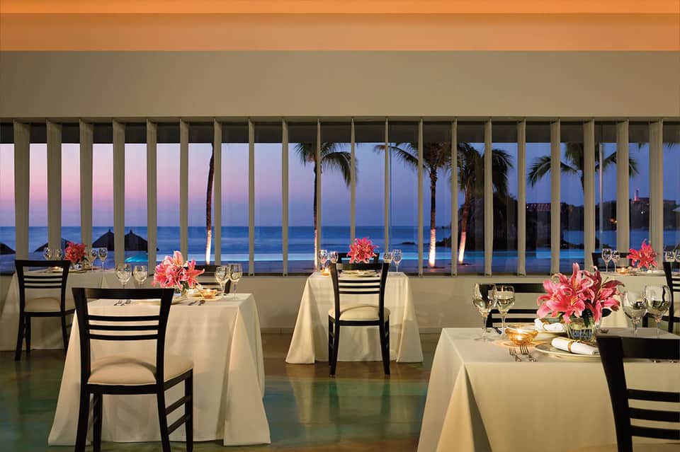 Fine dining restaurant with ocean views at dusk, elegant table settings and pink flowers